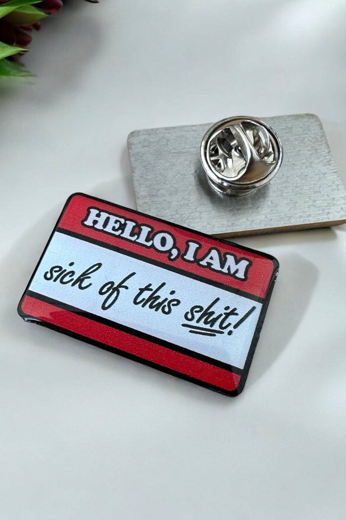 Eco emalje pin i rød/hvid med teksten Hello, I am sick of this shit, liggende på hvid baggrund ved siden af metal lås.