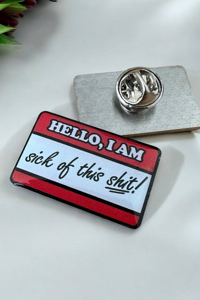 Eco emalje pin i rød/hvid med teksten Hello, I am sick of this shit, liggende på hvid baggrund ved siden af metal lås.