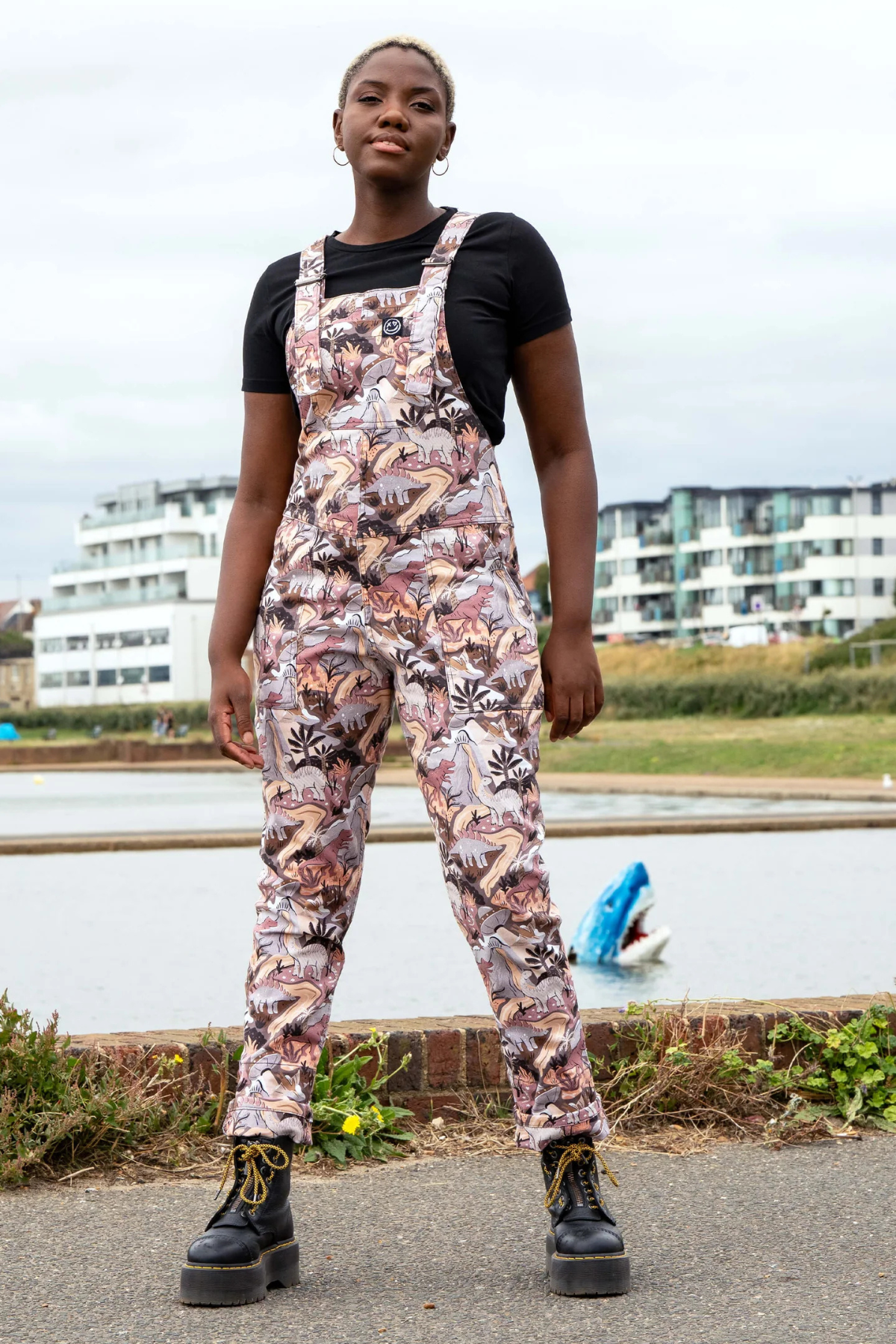 Model iført Rebel Romance Dinoverse Stretch Dungarees i støvede rosa og brunlige nuancer med dinosaur-print, stående foran vand og bygninger.