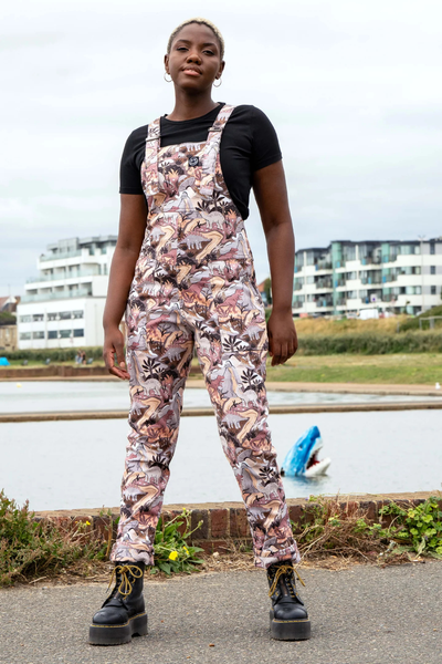 Model iført Rebel Romance Dinoverse Stretch Dungarees i støvede rosa og brunlige nuancer med dinosaur-print, stående foran vand og bygninger.