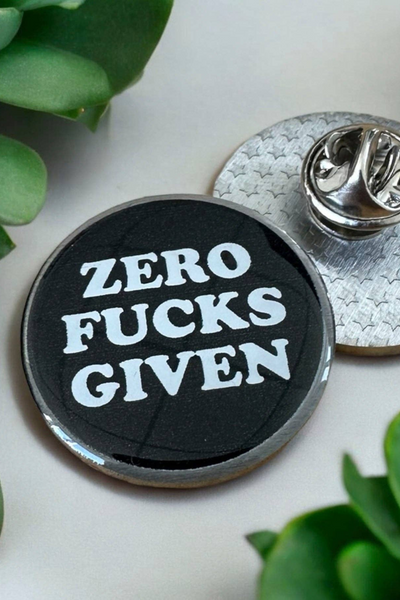 Rund sort Eco emalje pin med teksten Zero Fucks Given i hvid skrift, liggende på hvid baggrund ved siden af metal lås.