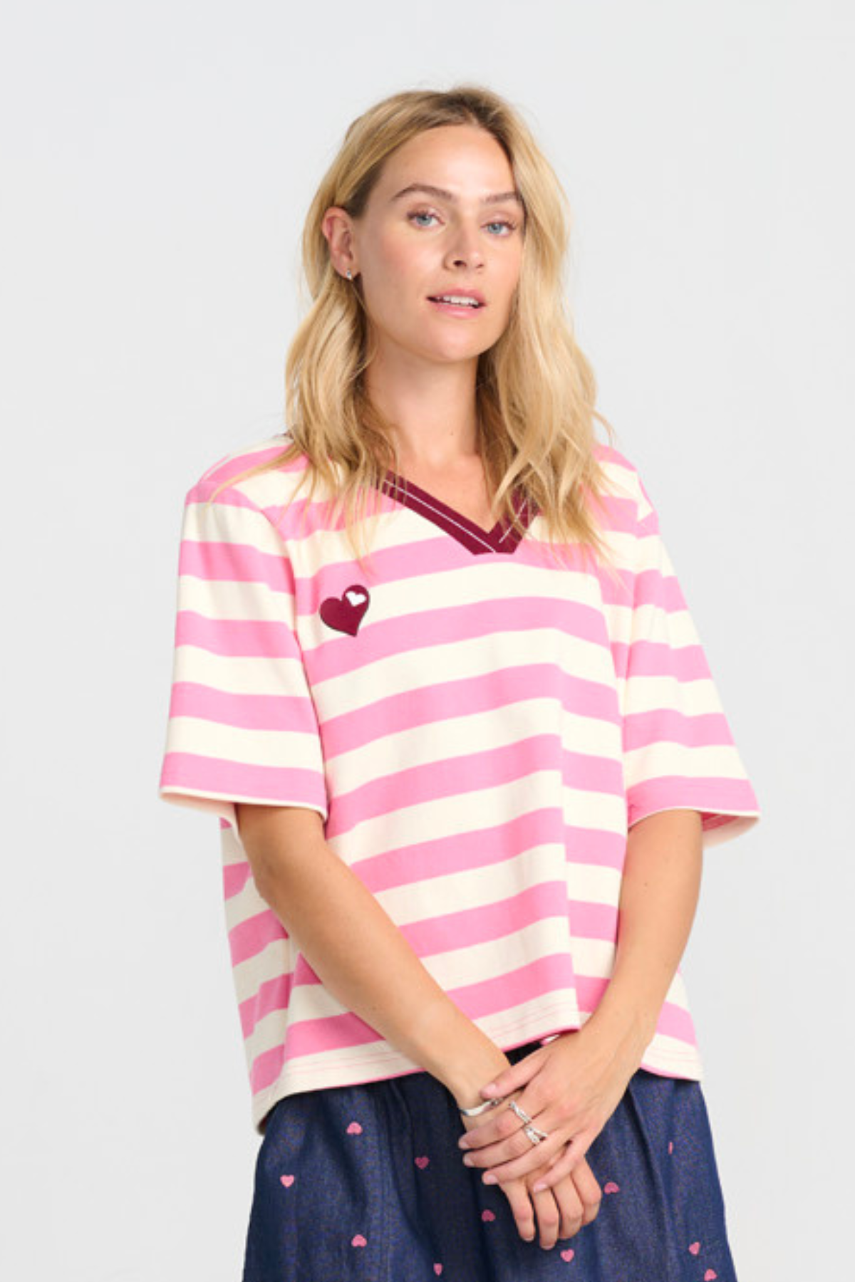 Nærbillede af modellen iført stribet tee i pink og offwhite med vinrød V-hals og lille hjerte på brystet