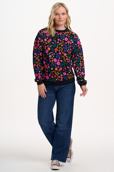 Blond model i farverig sweatshirt og jeans – fuld figur, afslappet look med sneakers.