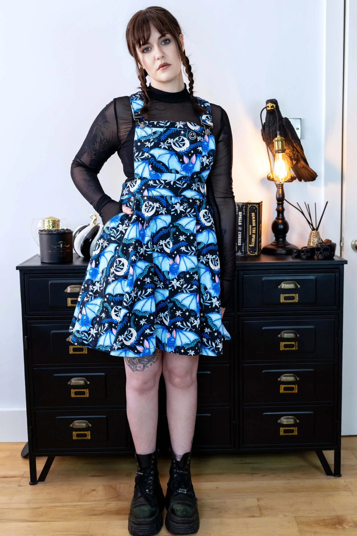 Rebel Romance Night Flight Pinafore vist på model med sort top og støvler, stående foran sort skuffemøbel.