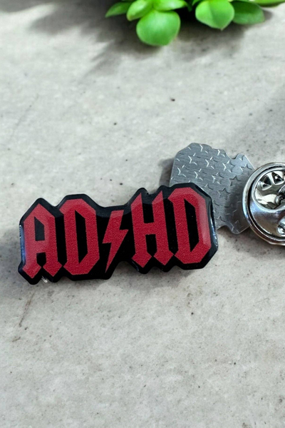Sort og rød Eco emalje pin med teksten ADHD og lyn-symbol, liggende på lys baggrund ved siden af metal lås.