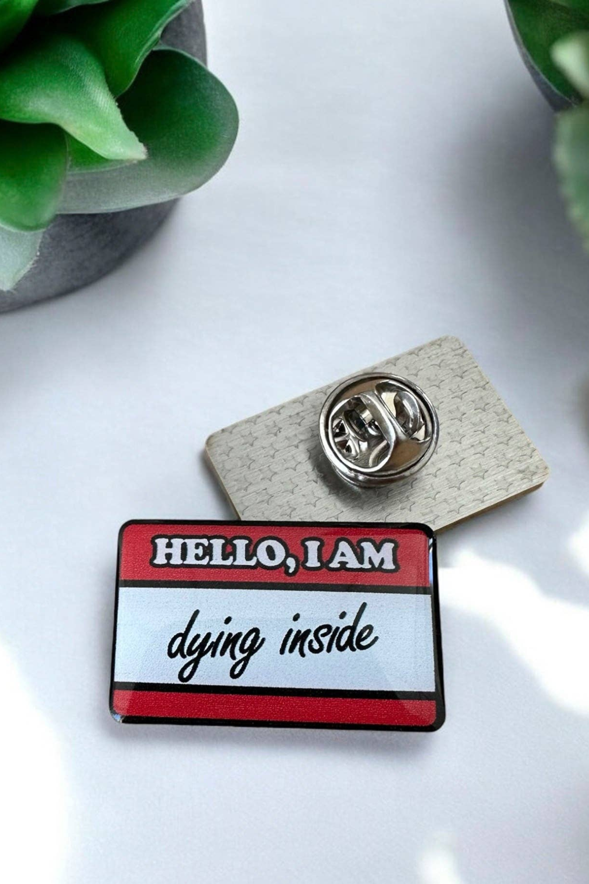 Eco emalje pin i rød/hvid med teksten Hello, I am dying inside, liggende ved siden af låsen på hvid baggrund med grøn plante.
