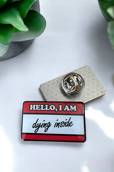 Eco emalje pin i rød/hvid med teksten Hello, I am dying inside, liggende ved siden af låsen på hvid baggrund med grøn plante.