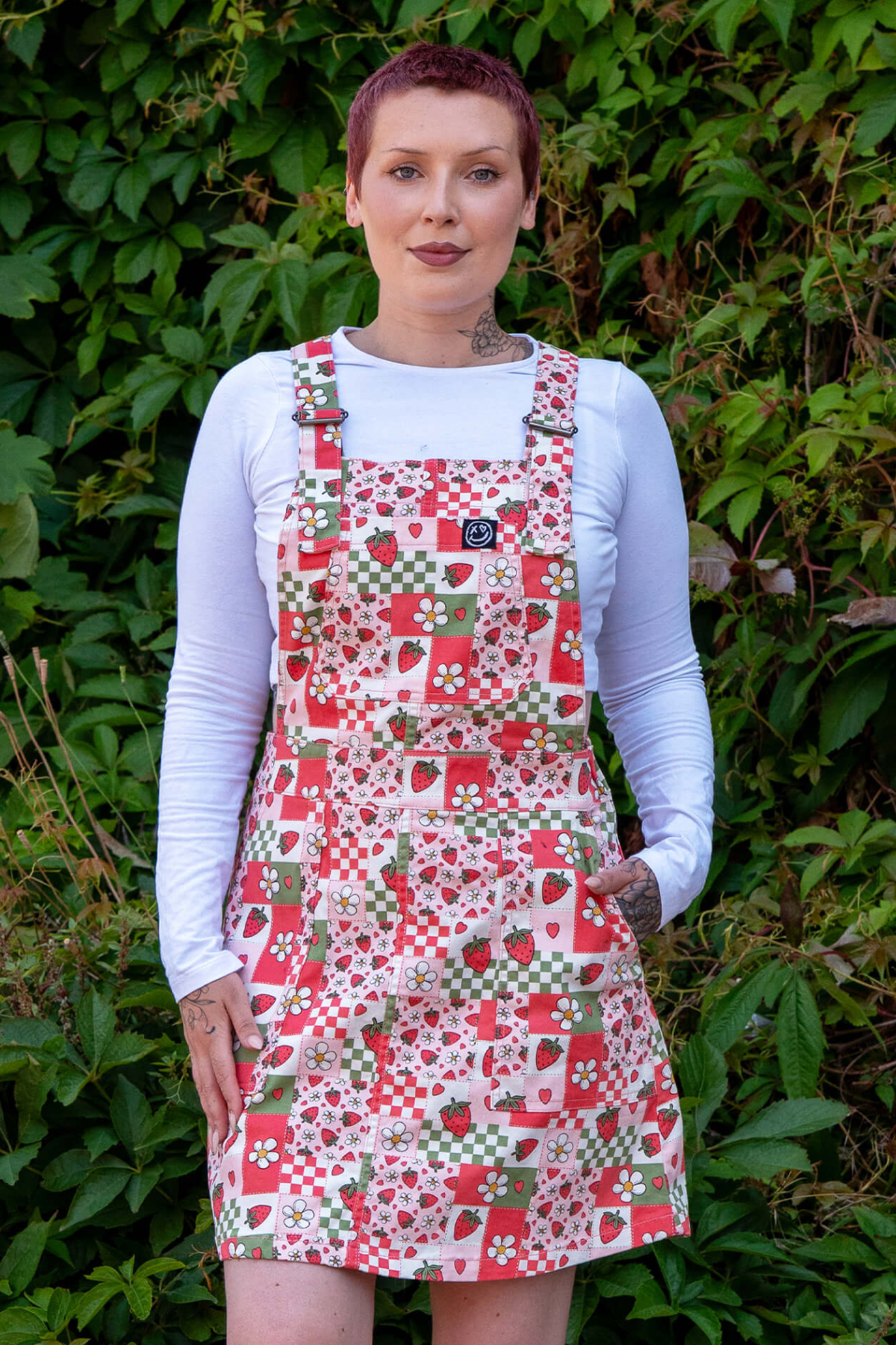 Katie iført Strawberry Picnic smæk-kjole og hvid langærmet top – står afslappet med hænderne i lommerne foran grøn baggrund