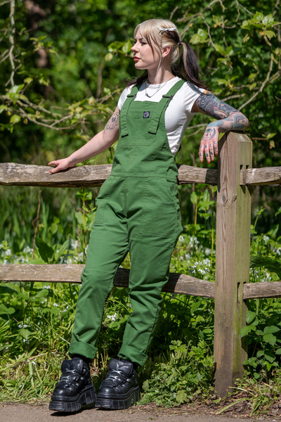 Florence iført Rebel Romance Forest Green Stretch Twill smækbukser, læner sig afslappet op ad et træhegn.