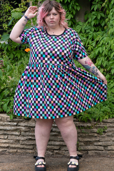 Sam iført Rebel Romance Checkerboard Smock Dress i en have med grøn baggrund, holder skørtet ud til siden