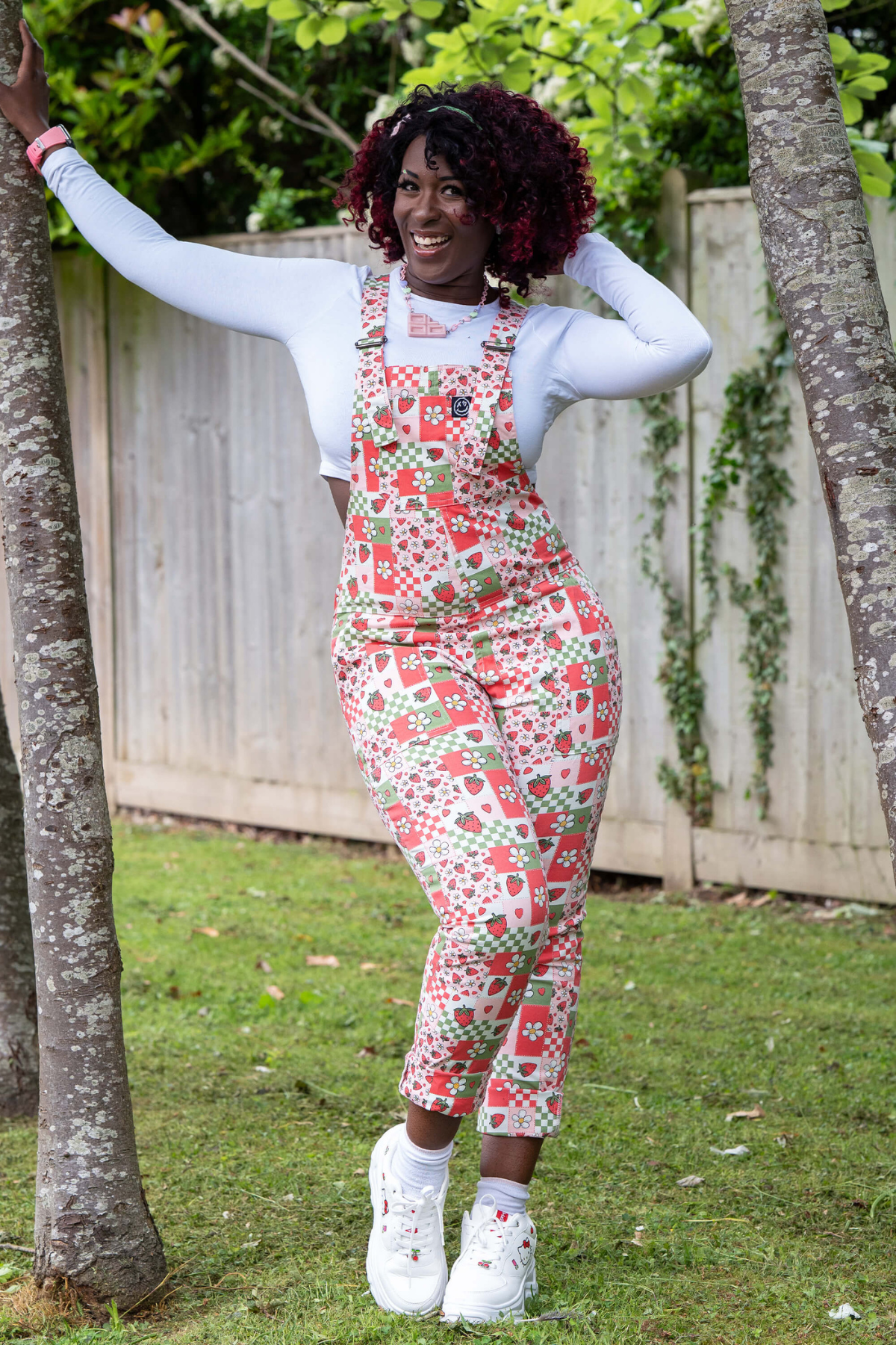 @ayeshashasha poserer i Strawberry Picnic Dungarees blandt træer, smilende og glad i farverigt print.