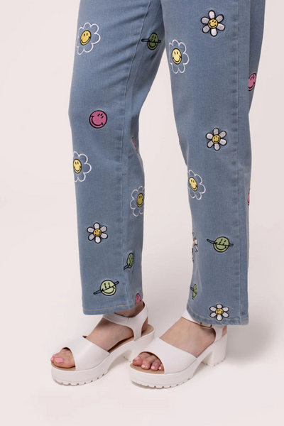 Nærbillede af benene i Lottie Jeans med broderede Smiley® blomster og planeter, stylet med hvide sandaler.