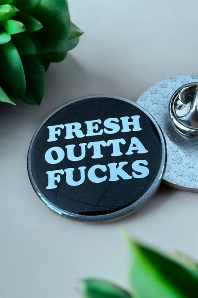 Rund sort Eco emalje pin med teksten Fresh Outta Fucks i hvid skrift, liggende ved siden af metal lås.