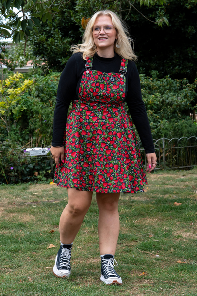 Rebel Romance Strawberry Field Pinafore med røde jordbær og grønne blade, vist på model i sort top og sneakers.