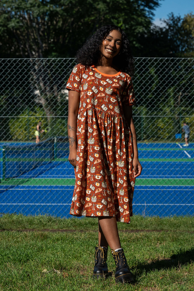 Model i Rebel Romance Mushroom Tea Party Dress med brun base og svampeprint, stående foran tennisbane.