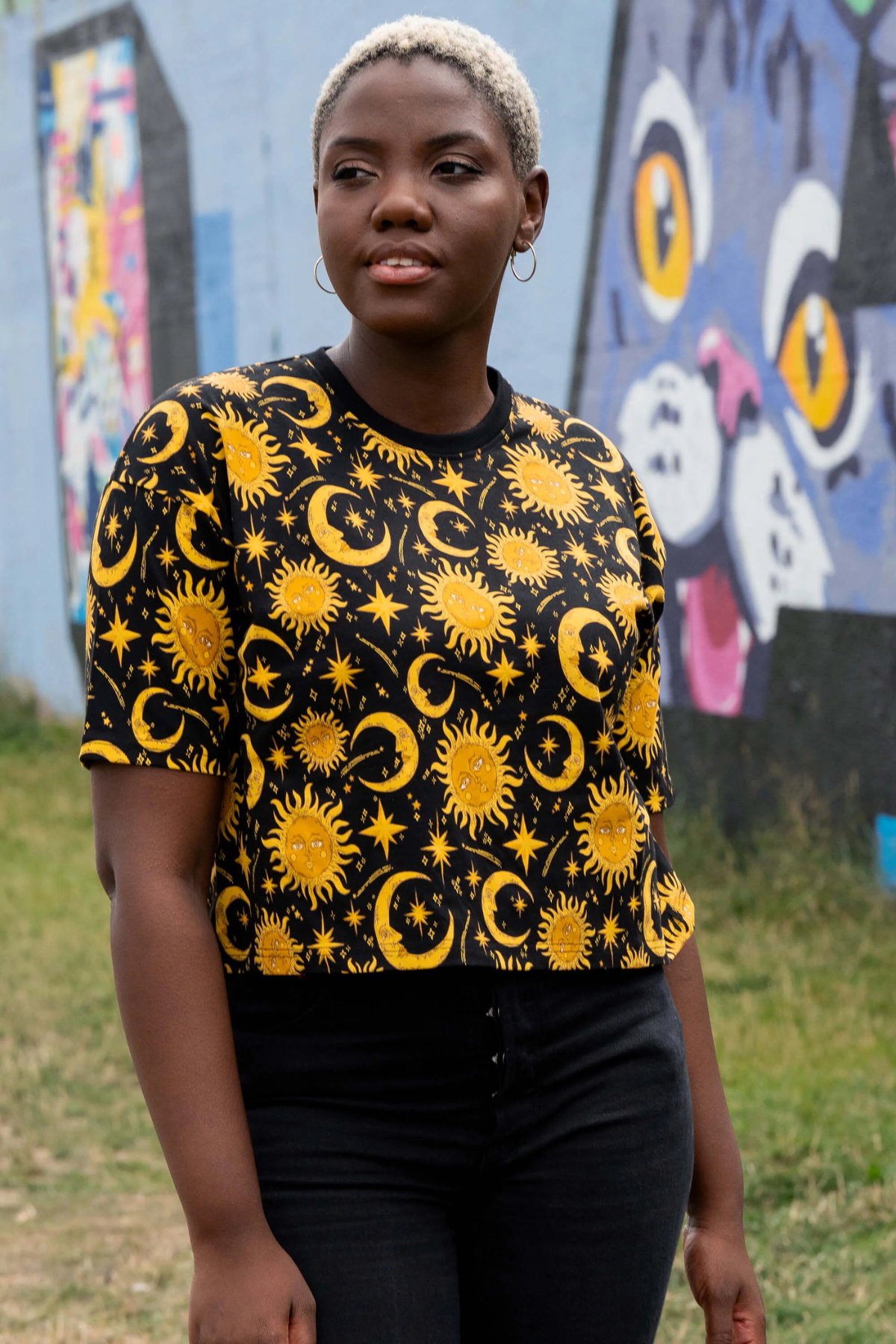 Model i Rebel Romance Celestial Sun & Moon Tee med sort base og gyldent sol- og måneprint foran farverig væg.