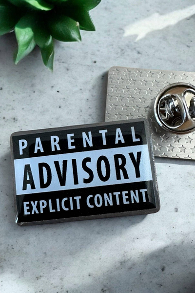 Eco emalje pin i sort/hvid med teksten Parental Advisory Explicit Content, liggende ved siden af metal lås på grå marmor baggrund.