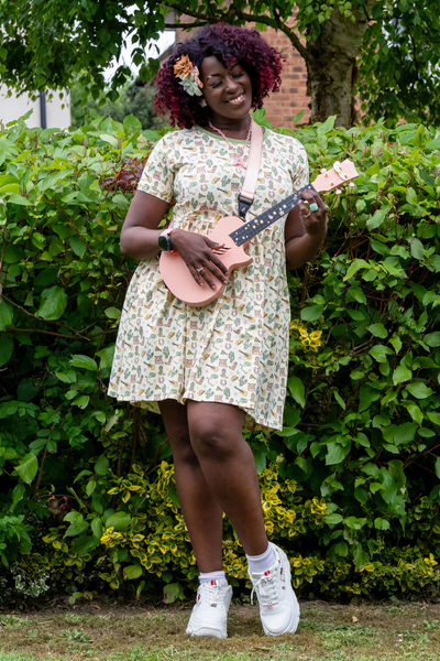 Ayesha smiler med ukulele i hånden iført smockkjolen – omgivet af grøn hæk