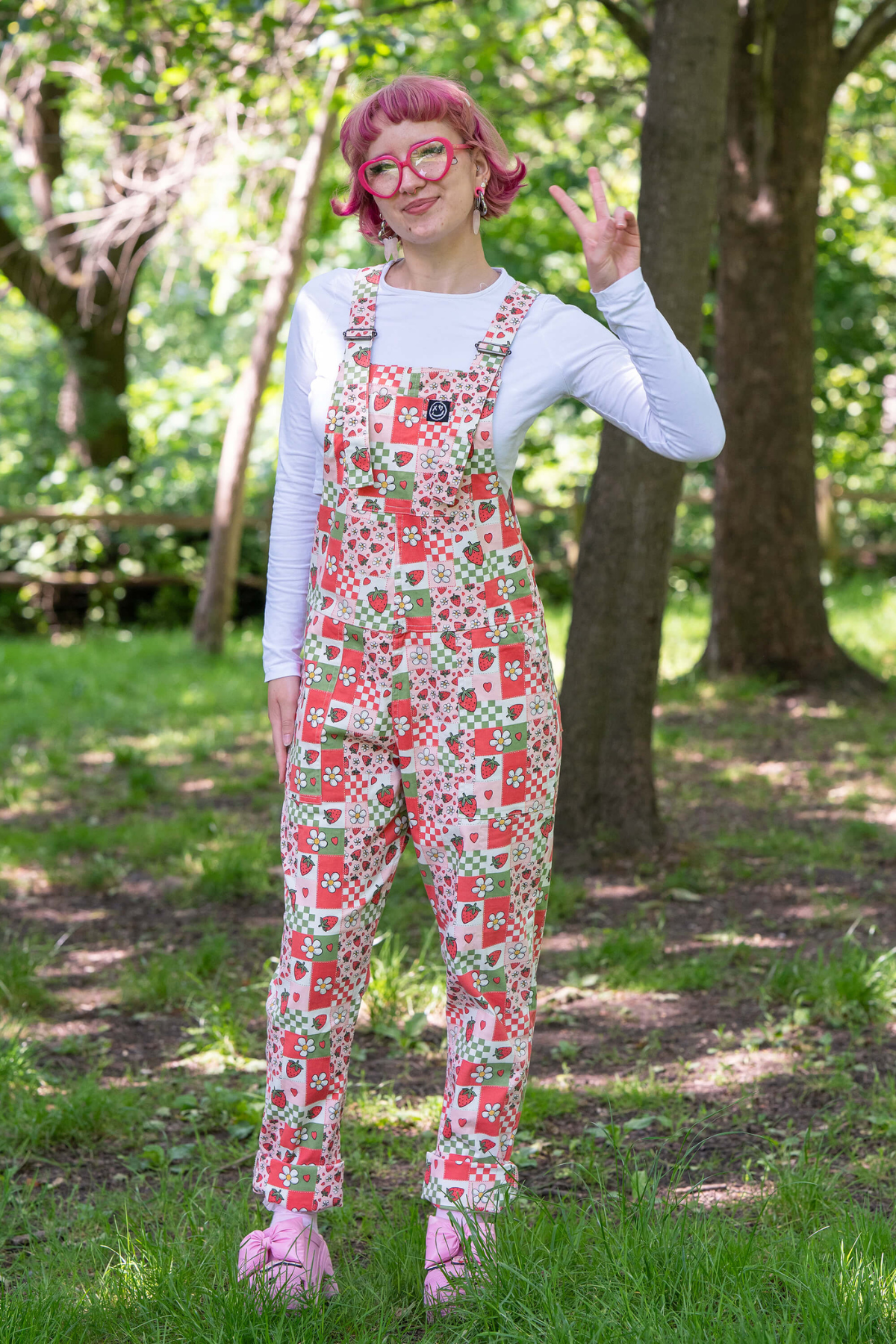 @fairyjesss står i sollys i Strawberry Picnic Dungarees, smilende og iført pink briller.