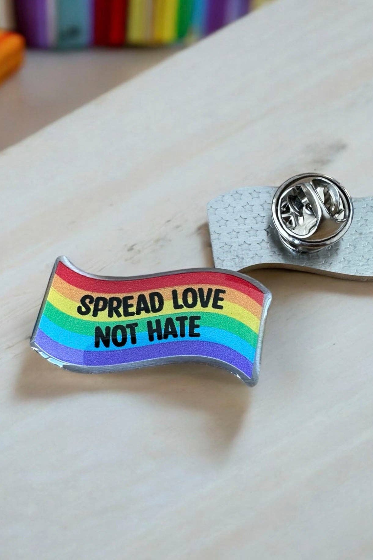 Regnbuefarvet Eco emalje pin med teksten Spread Love Not Hate, liggende på lys baggrund ved siden af metal lås.
