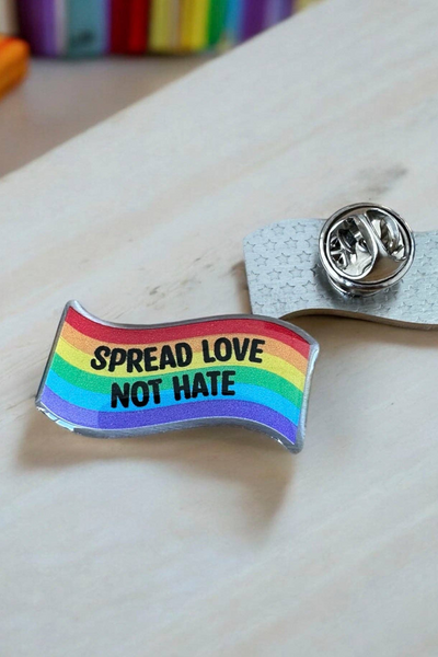 Regnbuefarvet Eco emalje pin med teksten Spread Love Not Hate, liggende på lys baggrund ved siden af metal lås.