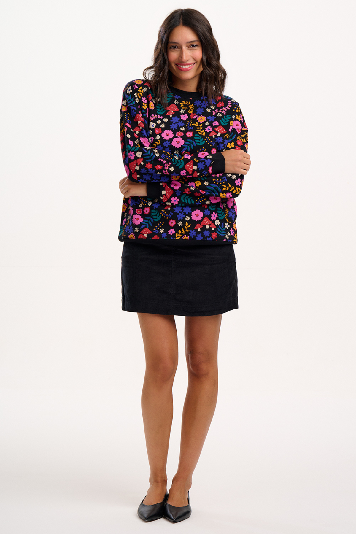 Model i farverig sweatshirt med blomstermønster og svampeprint – fuld figur, med sort nederdel og sorte sko.
