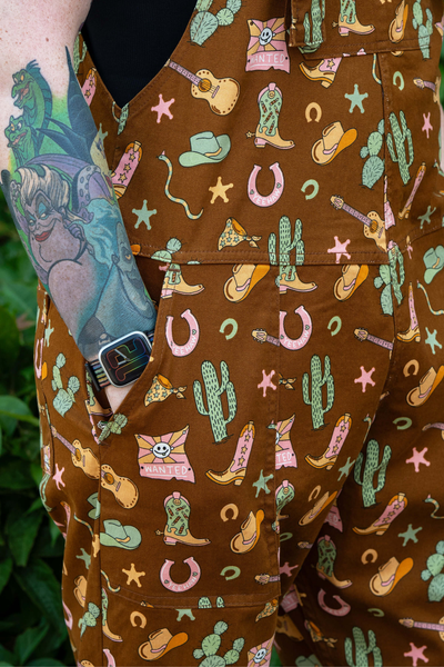 Detalje af print på Rebel Romance Cowboy Dungarees – kaktus, støvler, hatte og guitarer på brun baggrund.
