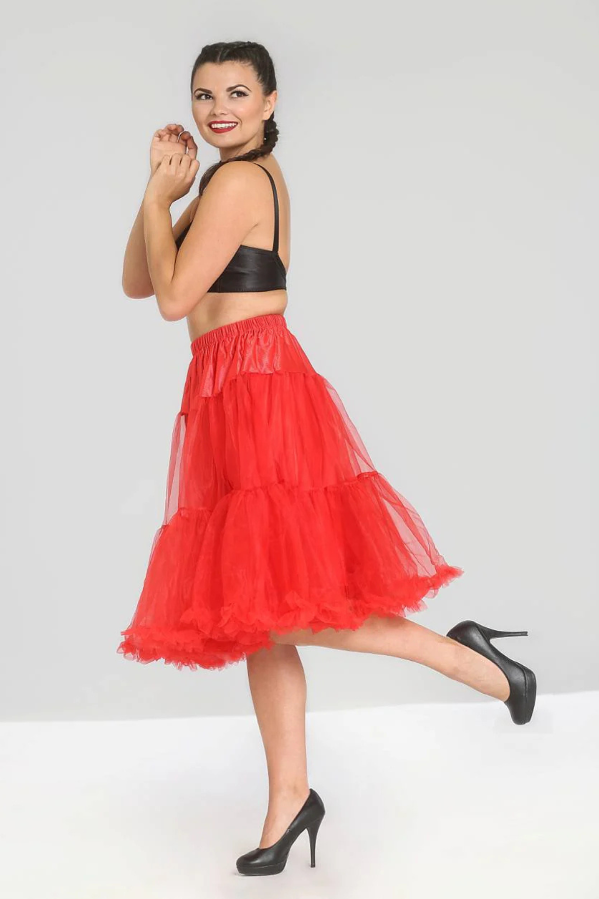 Model iført Hell Bunny Penelope petticoat i klar rød med masser af volumen og tyl, stylet med sorte stiletter og sort top – poserer legende i profil mod lys baggrund