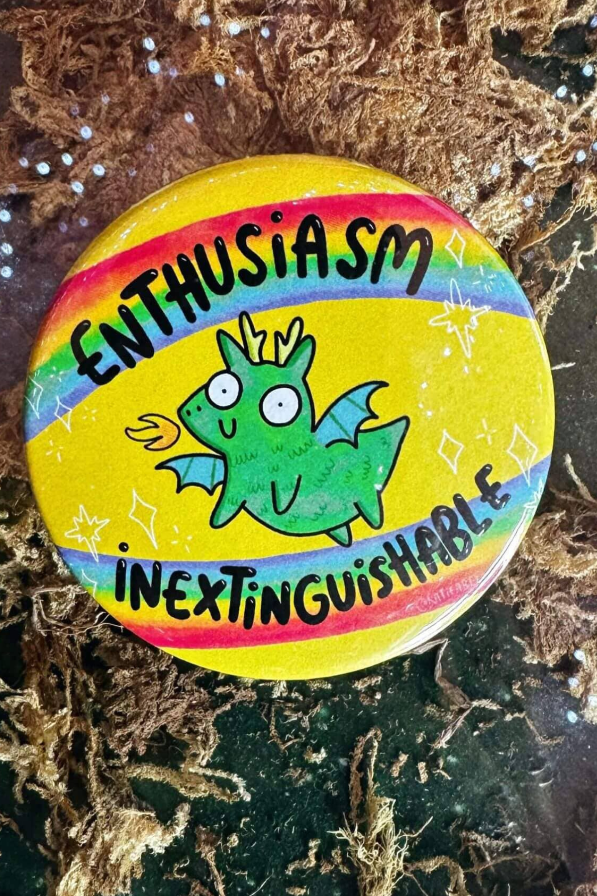 Katie Abey badge med teksten “Enthusiasm Inextinguishable” og en lille grøn ildspyende drage på regnbuebund – 58 mm badge