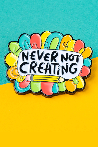 Farverig “Never Not Creating” emaljepin med regnbuemotiv på turkis og gul baggrund