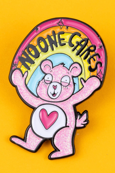 Katie Abey “No-One-Cares Bear” pin – glittery lyserød bjørn med regnbue og teksten No One Cares, soft enamel, på gul baggrund