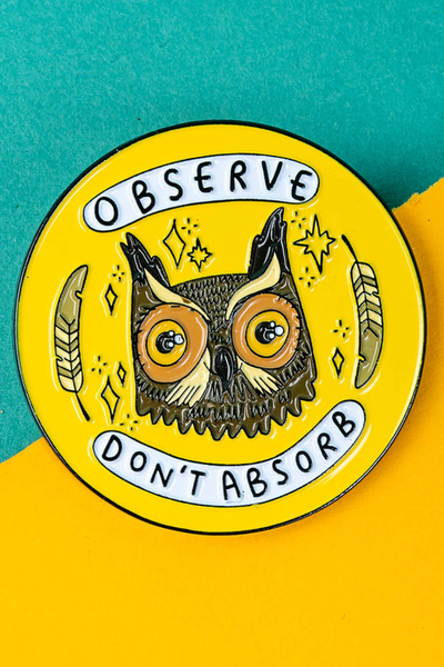 Gul rund “Observe – Don’t Absorb” emaljepin med uglemotiv på farverig baggrund