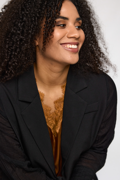 Model smiler iført kobberfarvet Ingrid top fra STUDIO under sort blazer.
